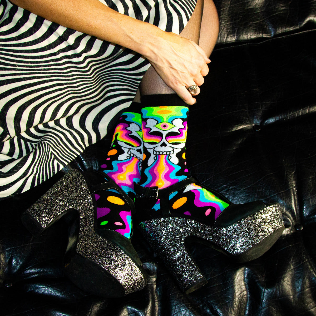 A person wears sparkly black platform heels with crew socks with psychedelic rainbow skulls and a black and white print skirt