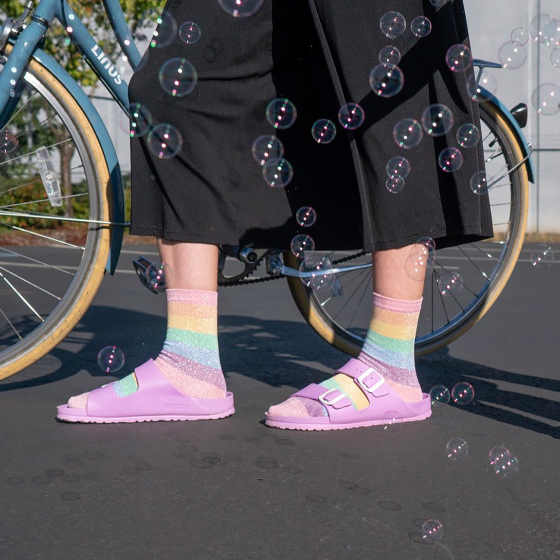 A person wears pastel rainbow crew socks with pale purple sandals and a black skirt while soap bubbles swirl around