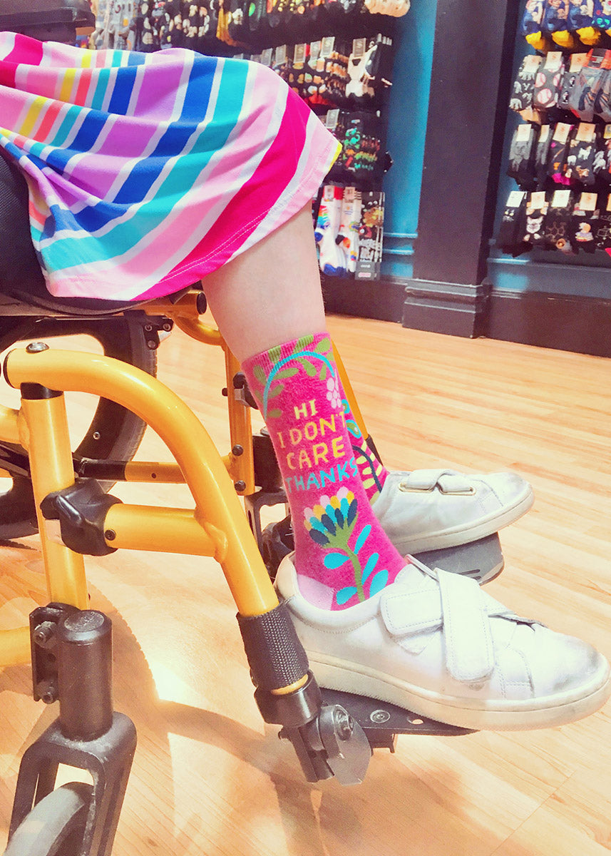 A person wears pink socks with a funny saying with white shoes and a rainbow dress while using a yellow wheelchair