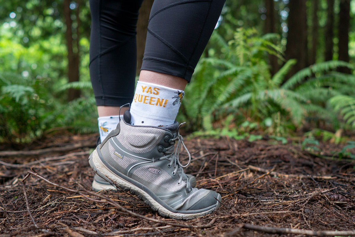Cuffed ankle socks that say "Yas Queen" worn with athletic shoes and leggings on a forested trail.