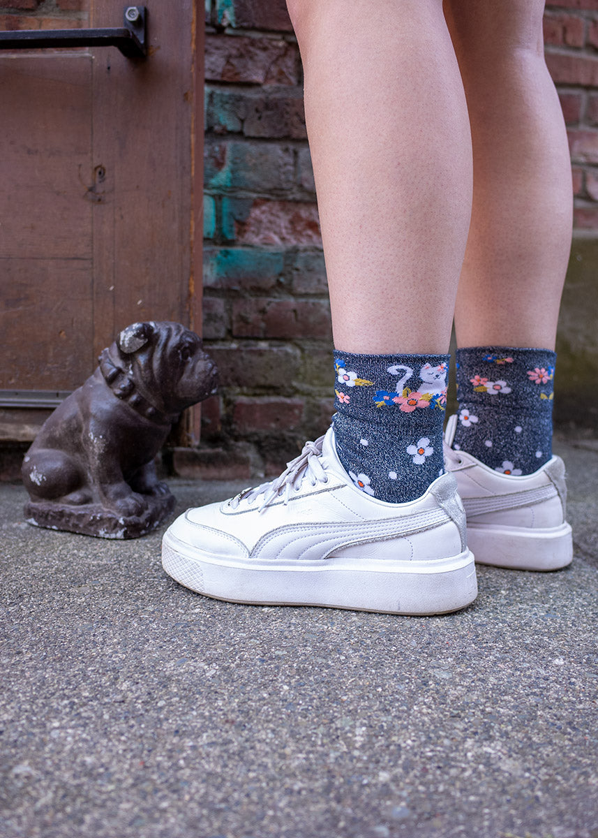 Sparkly cuffed ankle socks with cats on them are worn with white sneakers next to a small sculpture of a bulldog