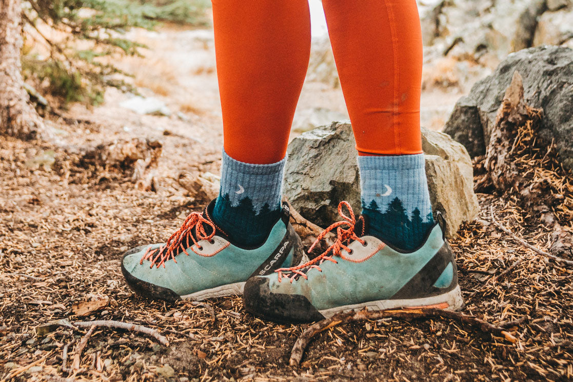Blue merino wool hiking socks with trees are worn on a trail with red leggings and teal shoes