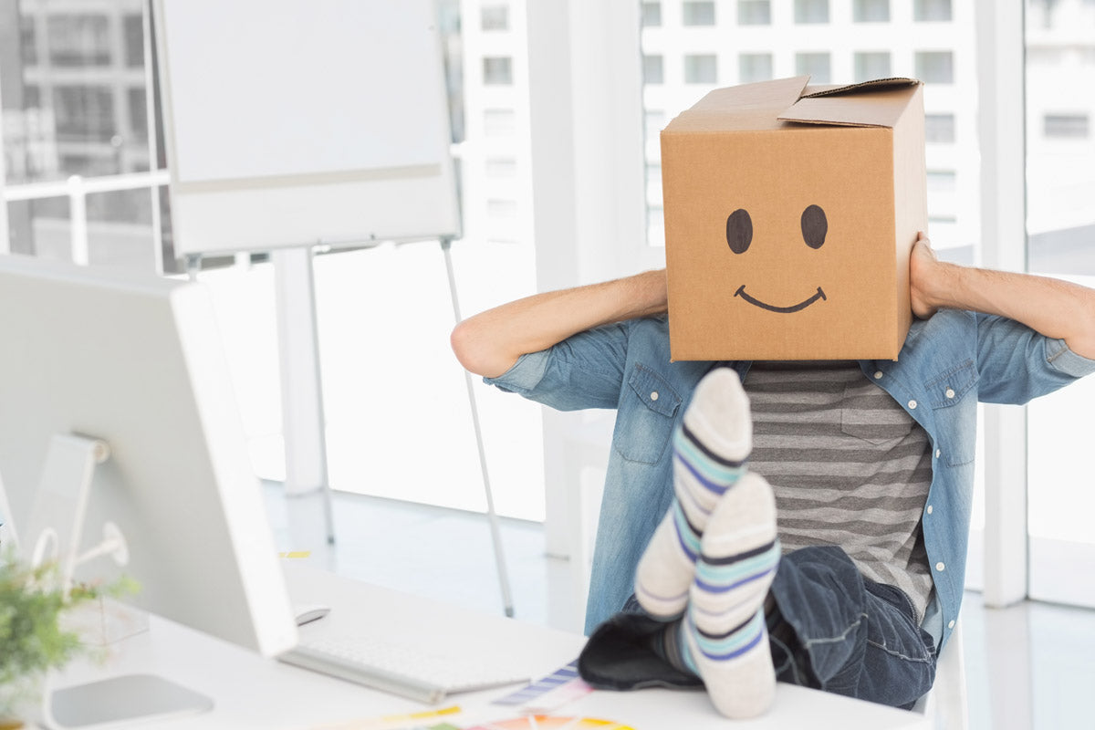 A man in striped sock kicks his feet up on his desk while wearing a cardboard box with a smiley face in a relaxed pose