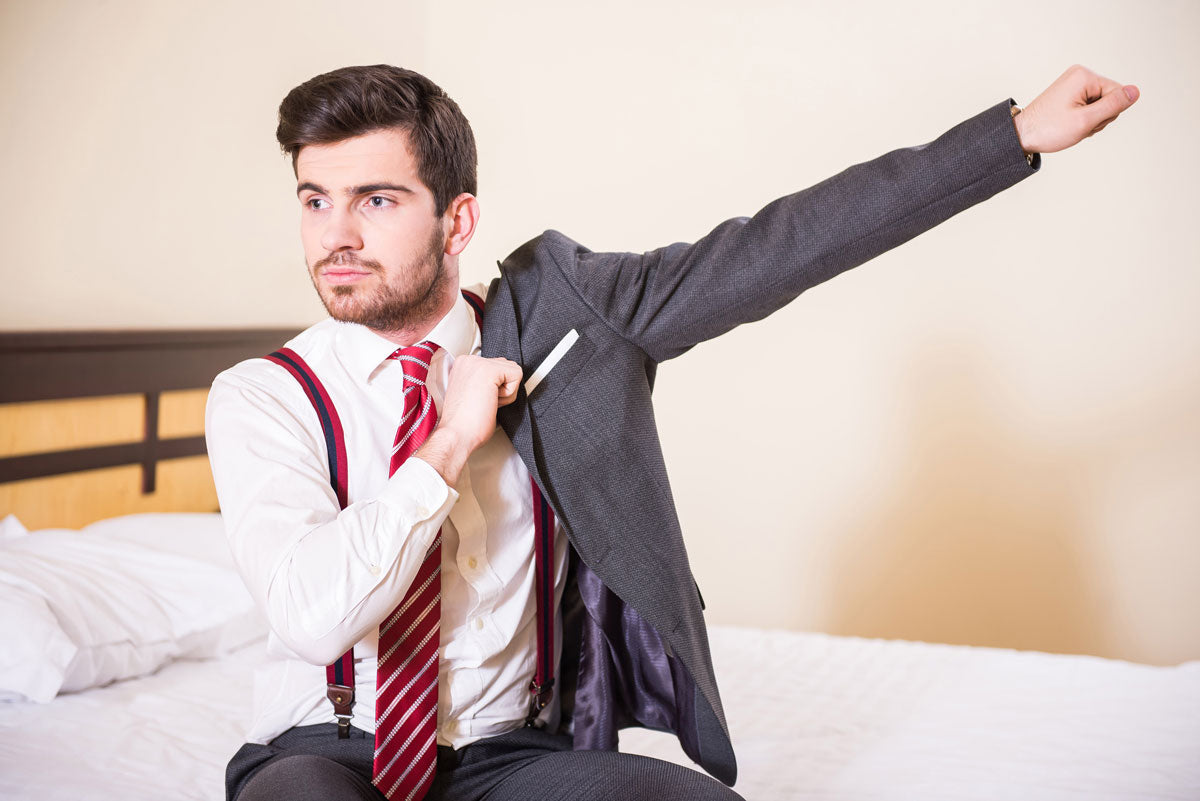 A man in business dress pulls on the arm of a suit jacket while sitting on a bed