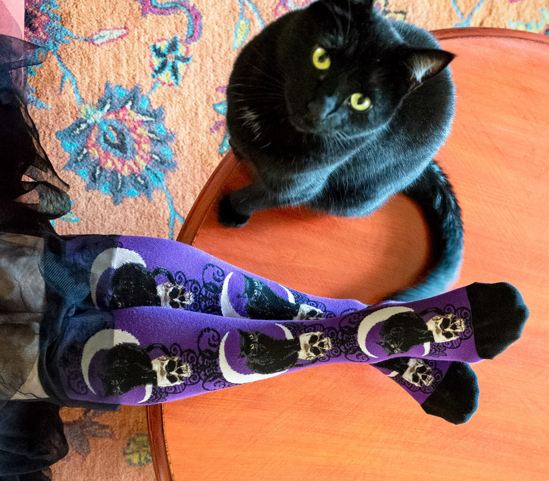 Purple knee socks with cats, skulls and moons sit beside an actual black cat
