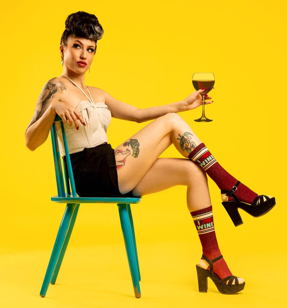 A woman sits in a chair and holds a glass of wine while wearing socks that say "I Heart Wine"