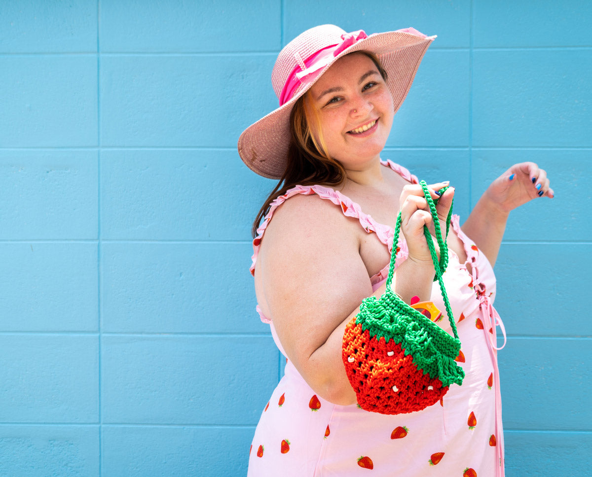 A woman in a pink hat and dress holds a purse shaped like a strawberry