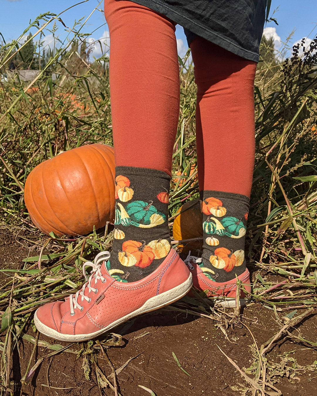 Andrea wears Oh My Gourd Socks by ModSocks with orange shoes and stands in a pumpkin patch