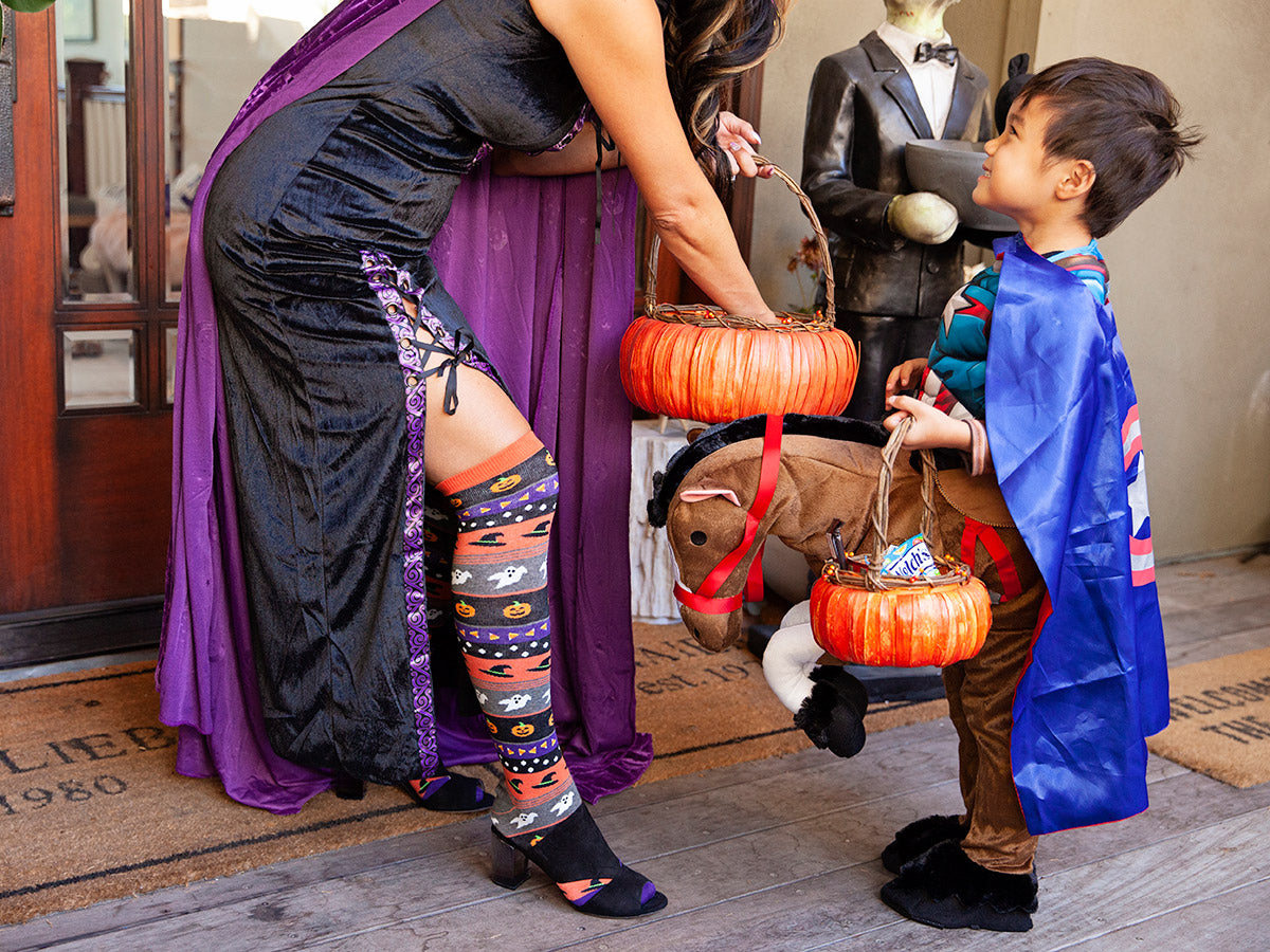 A trick-or-treater is offered candy by a witch wearing Halloween knee-high socks available at Cute But Crazy Socks
