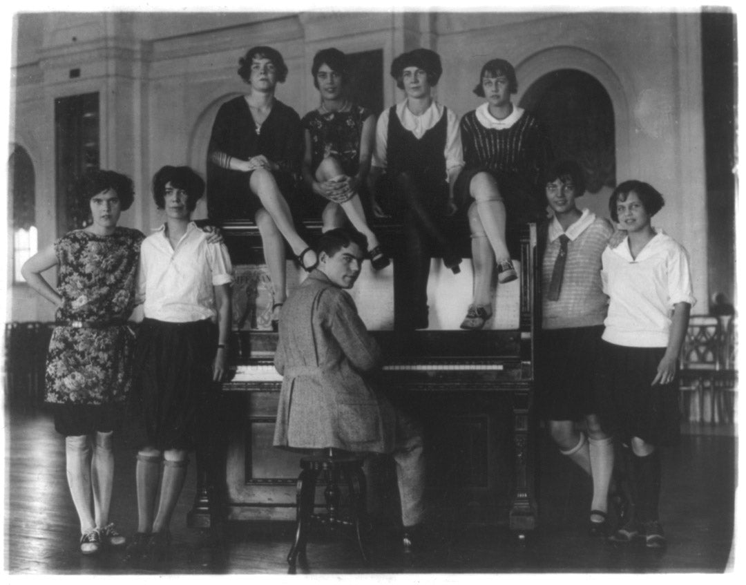 Young women "flappers" surround a piano player in the 1920s. Many of the women wear their stockings rolled down past their knees.