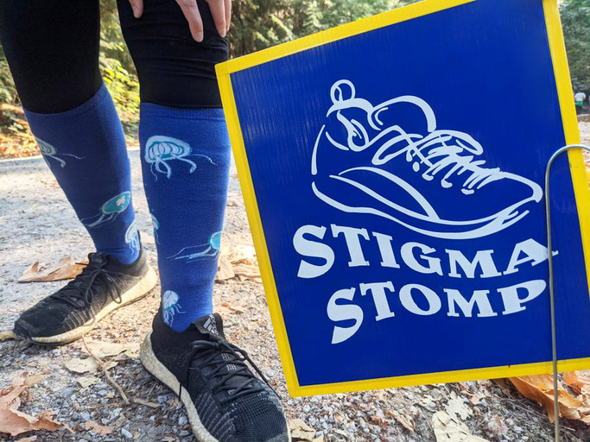 A person wearing blue jellyfish knee socks stands beside a blue sign reading "Stigma Stomp"
