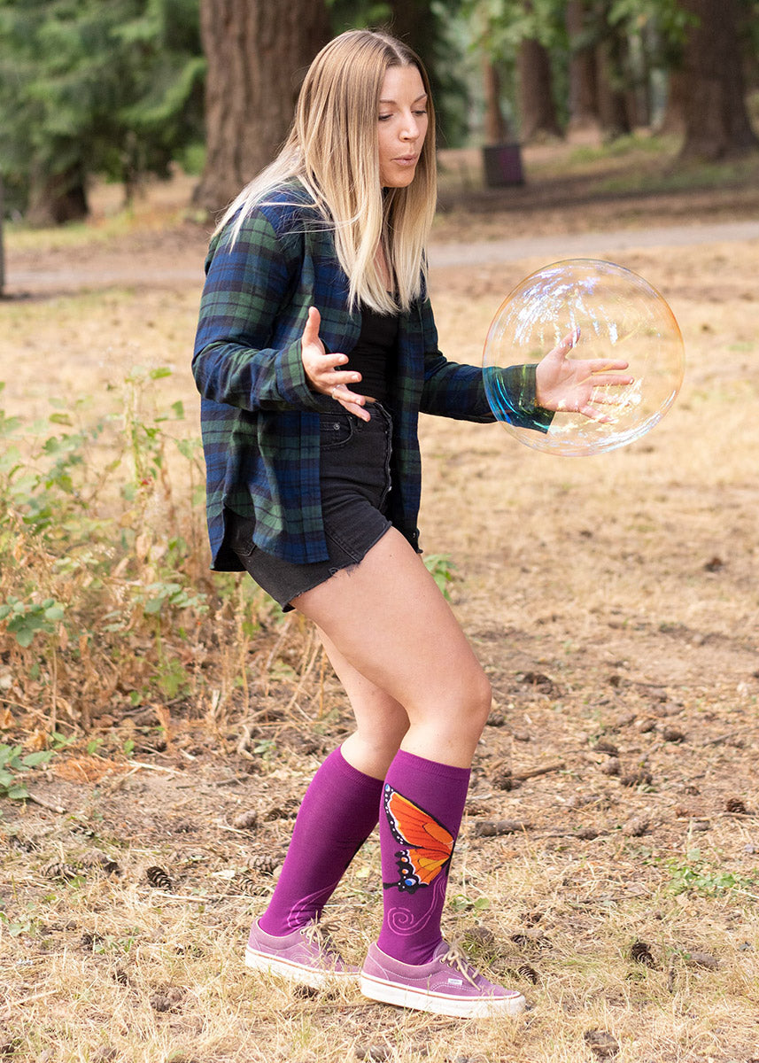 A woman wears purple socks with butterfly wings along with shorts and a plaid shirt while standing outside next to a giant soap bubble