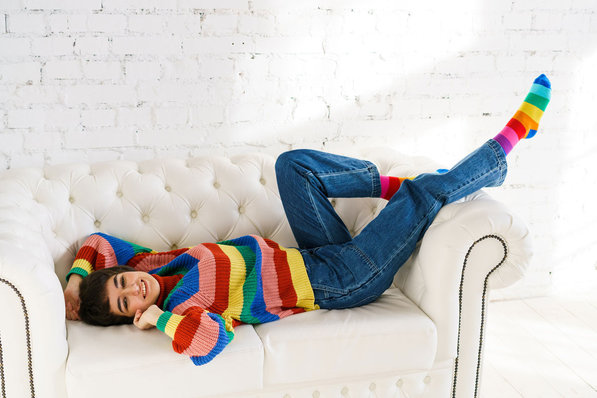 A woman lays on a couch with her feet up in rainbow stripe knee socks, jeans and a rainbow sweater