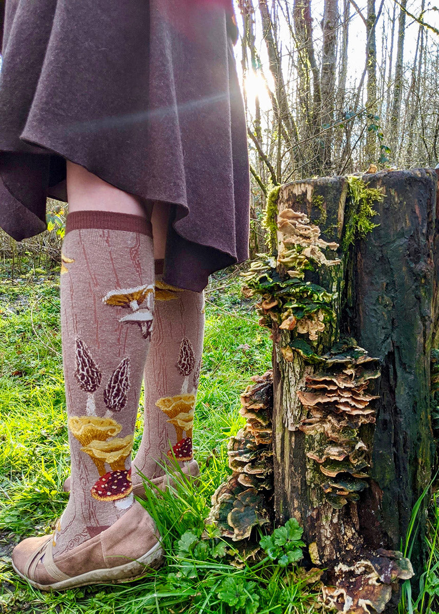 A person stands outdoors in mushroom knee socks next to a fungus-covered stump
