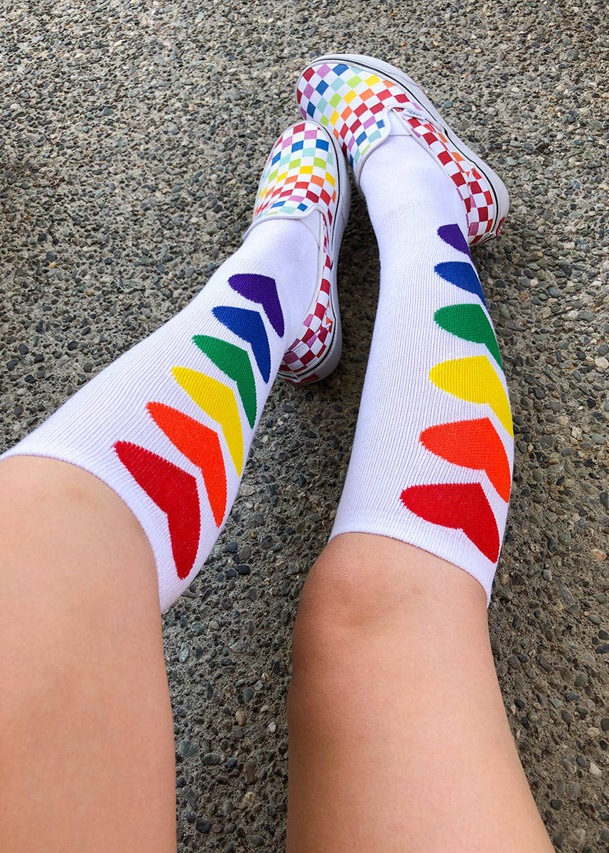 Fun knee socks with rainbow hearts on a white background are worn with rainbow checkerboard shoes.