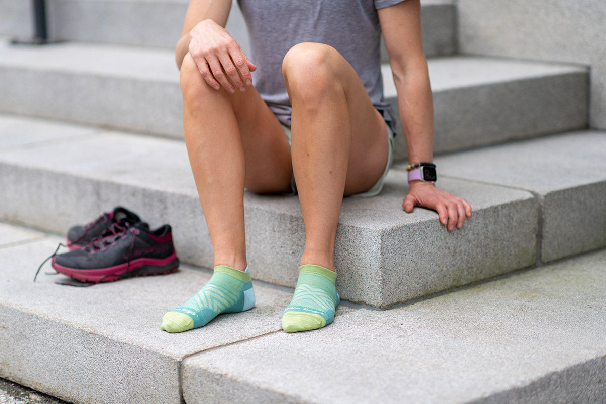 A person in aqua blue low-cut ankle socks sits on steps beside a pair of running shoes