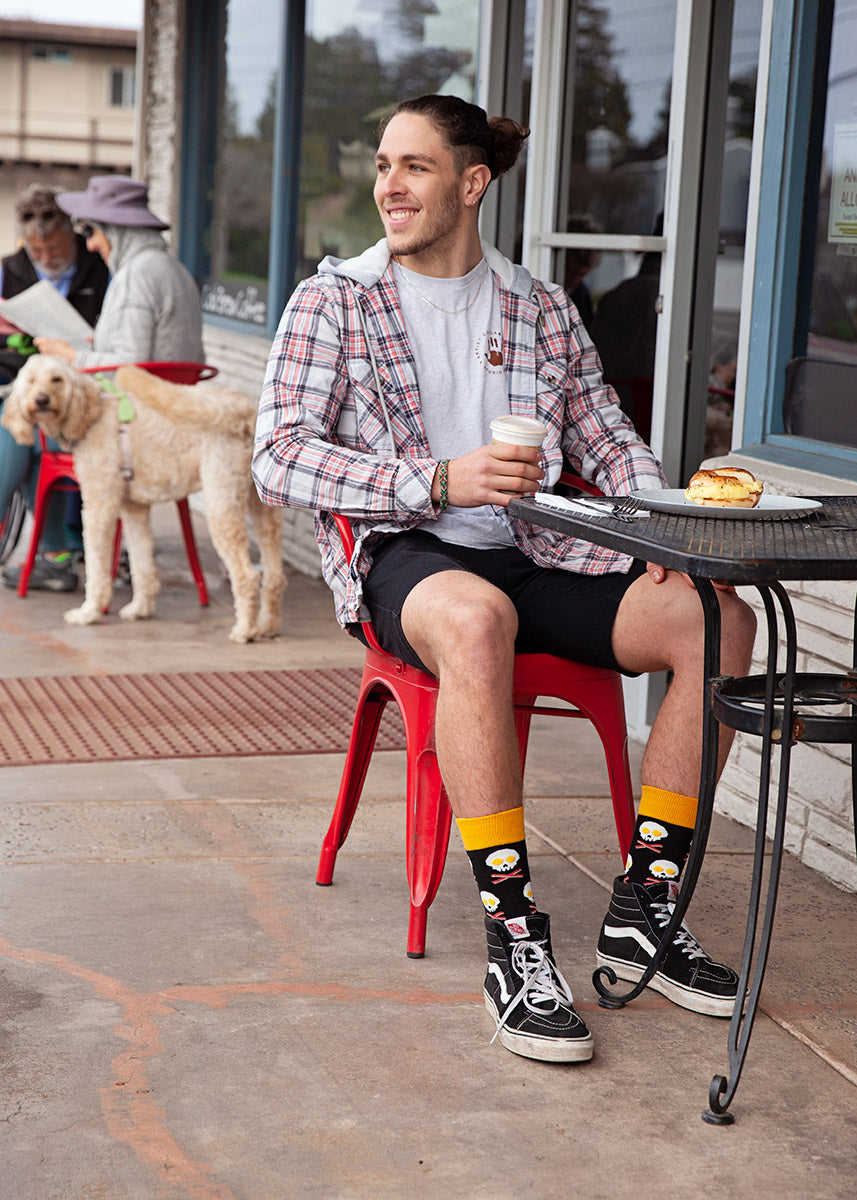 A man sits at an outdoor restaurant table wearing shorts and socks with bacon and egg skulls.