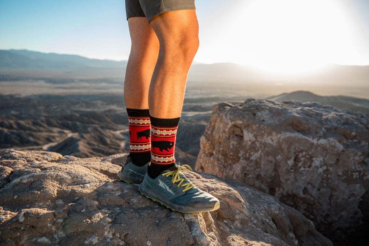 A man stands on a rock wearing shorts, sneakers and hiking socks with buffalo on them.