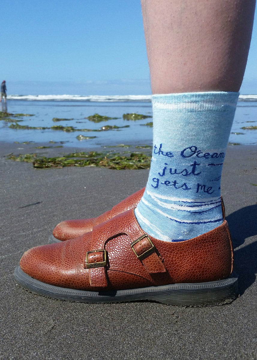 Socks that say "The Ocean Just Gets Me" standing on a beach