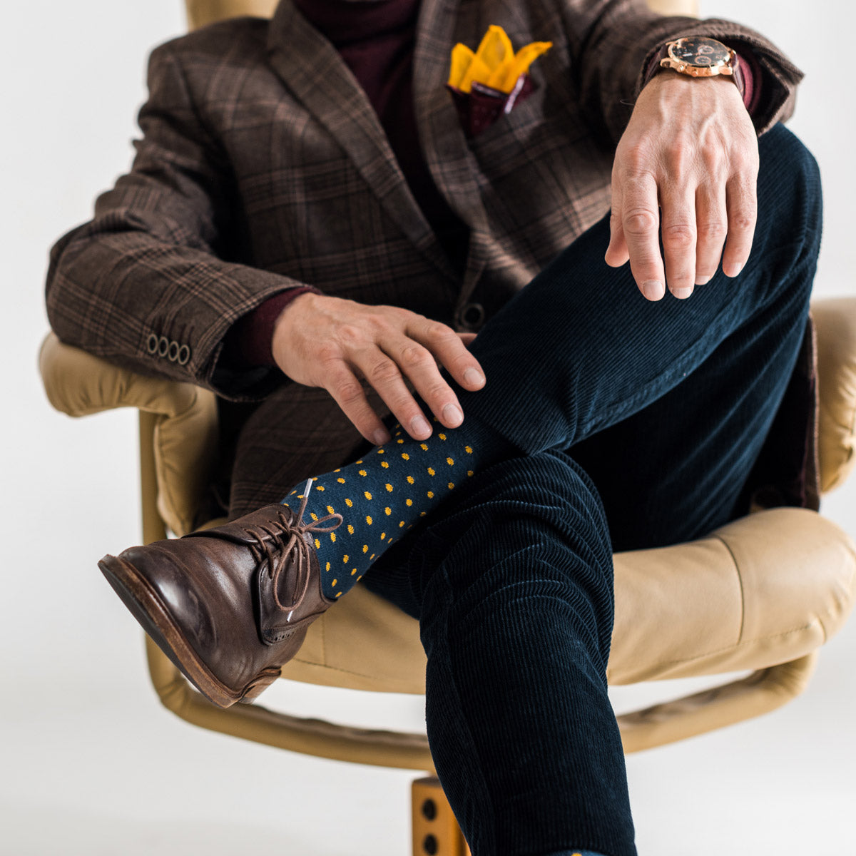 A man crosses his legs to show dress socks in navy with yellow polka dots worn with brown men's dress shoes