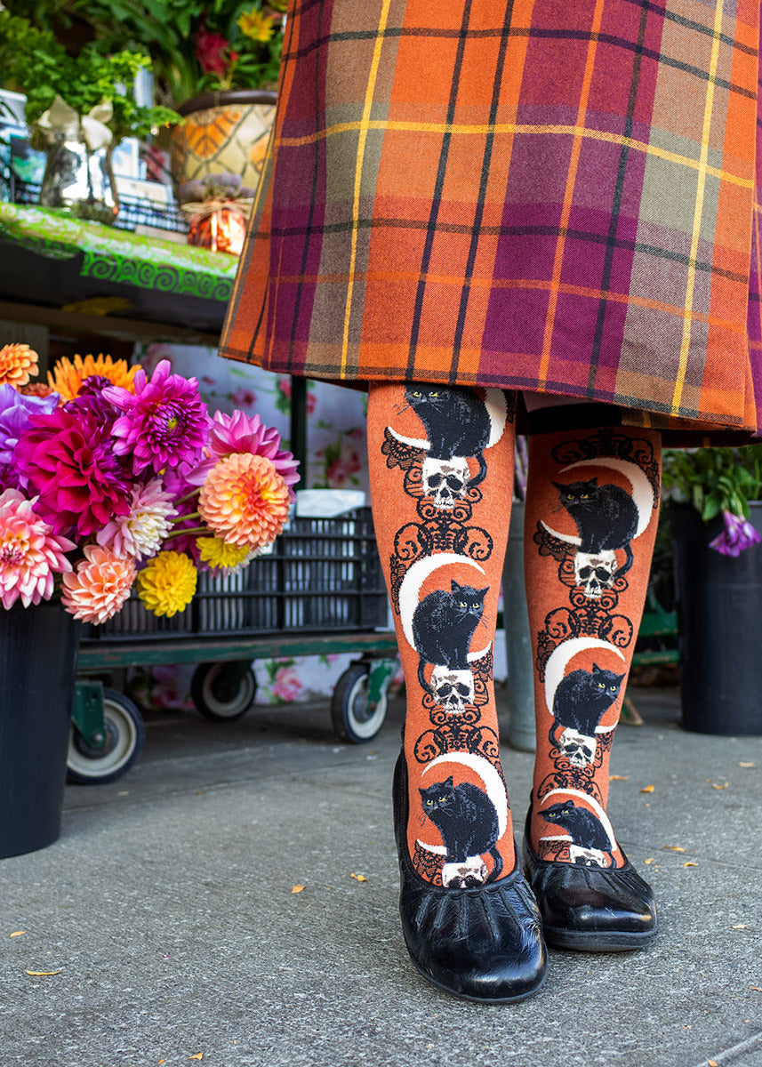 Black Cat Moon Knee Socks by ModSocks worn with plaid skirt beside a flower display