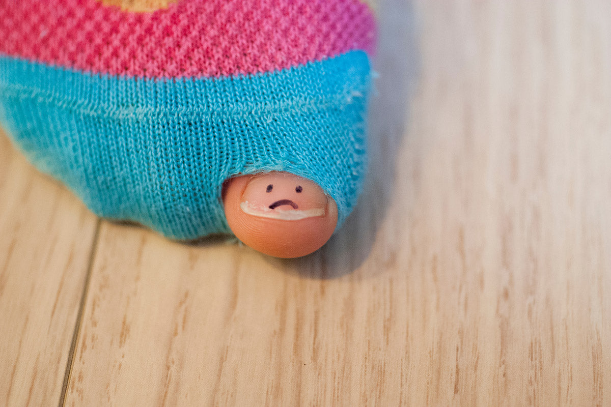 A big toe with a sad face drawn on the toenail peeks through a hole in a worn pair of pink and blue socks