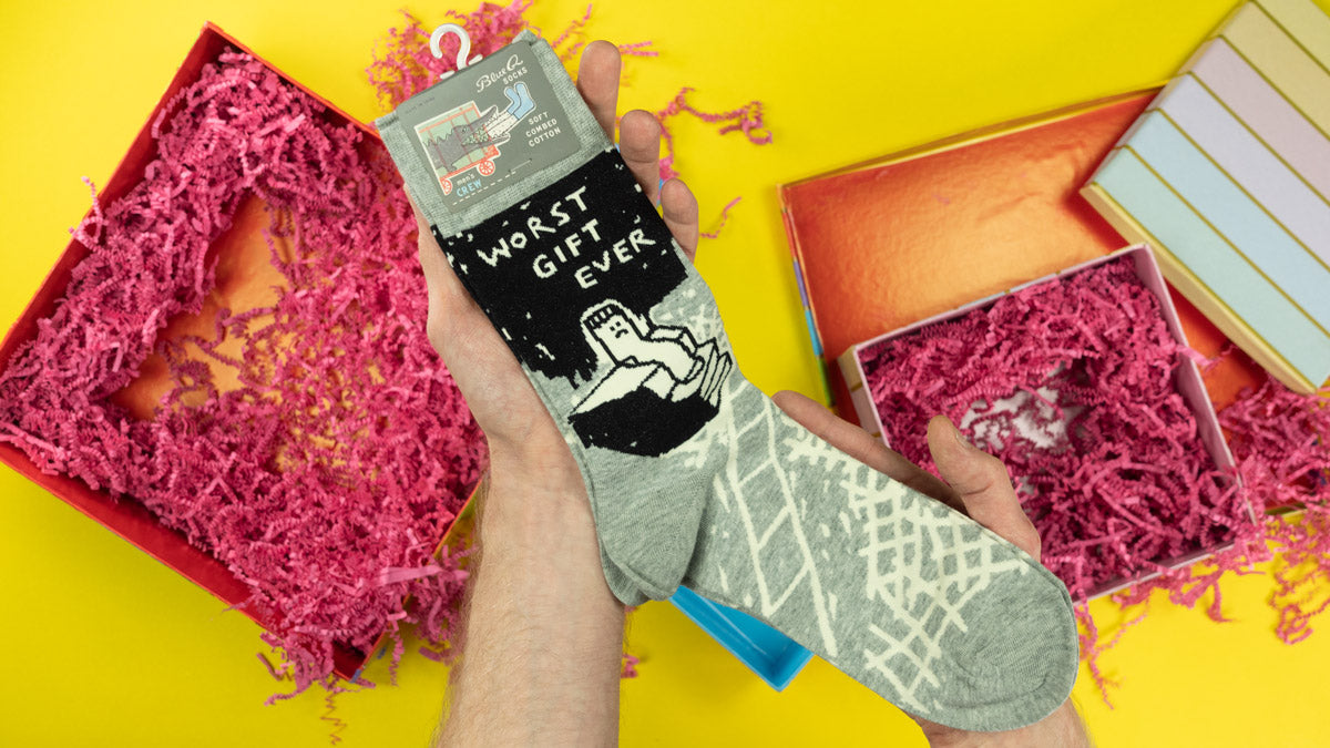 A person holds funny socks that say "Worst Gift Ever" above gift boxes with shredded crinkle paper filling