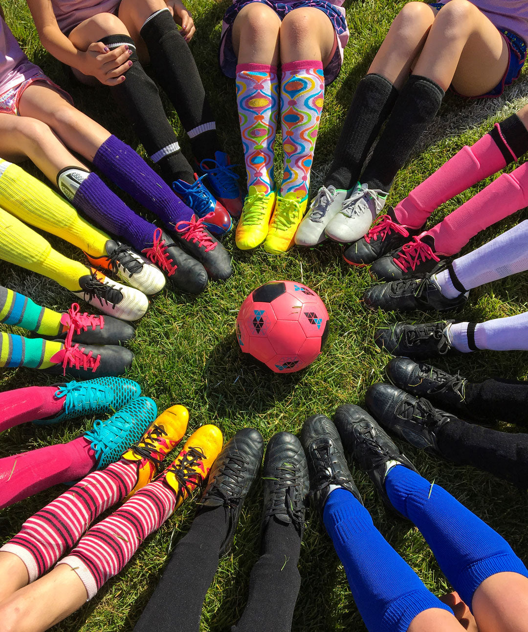 Soccer players show off their colorful socks sitting in a circle around a pink soccer ball.