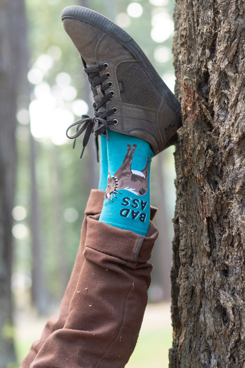 A man poses upside down against a tree trunk in donkey socks that say "Bad Ass"
