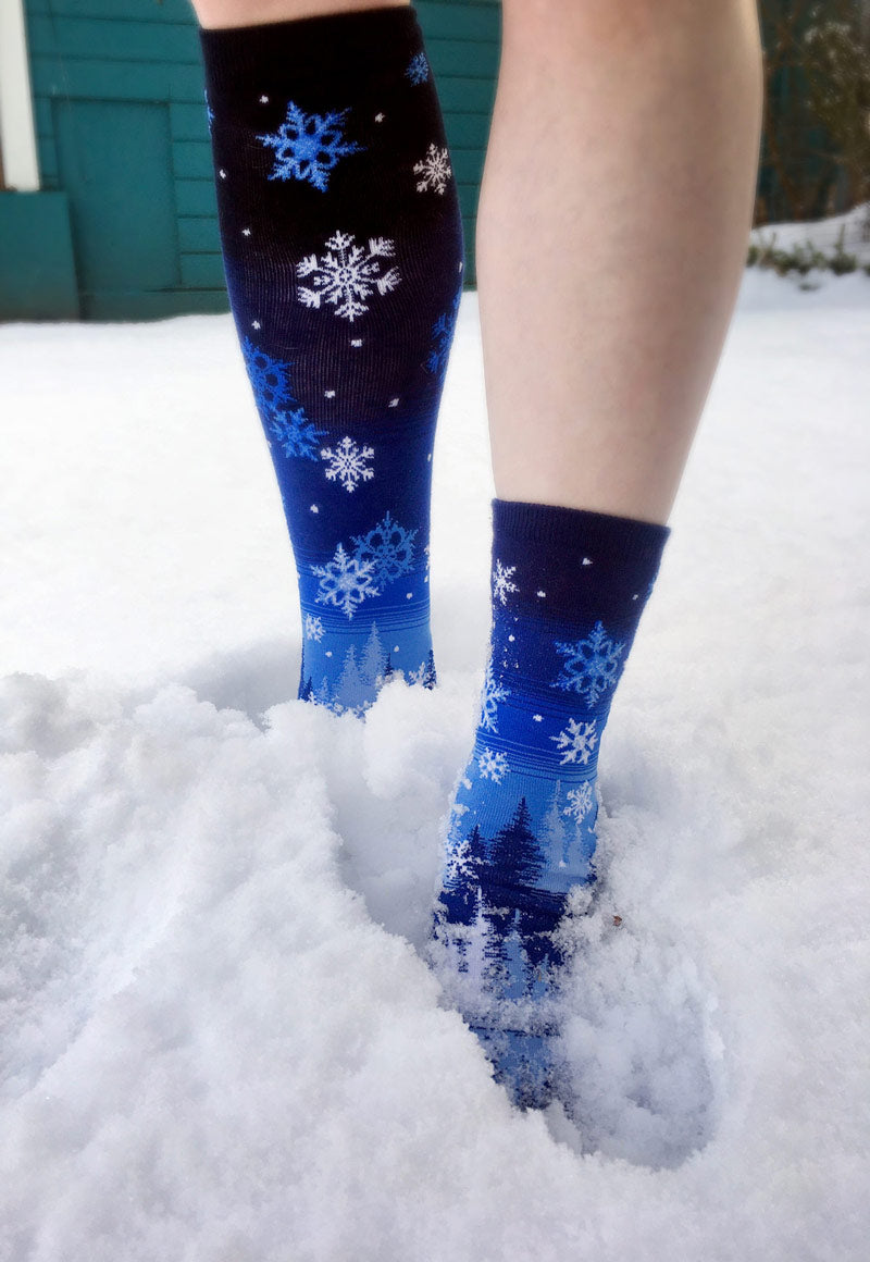 Someone stands in the snow wearing snowflake socks with no shoes.
