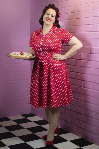 Dark haired woman standing in a 50s style diner holding a dessert on a platter and wearing a red and white polka dot dress with red shoes