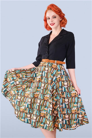 Red haired woman swishing her skirt and smiling. She is wearing a black blouse and a 50s style book print swing skirt.