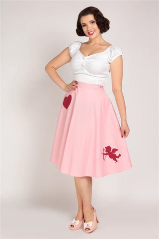 Smiling woman standing with on hand on her hip wearing a pink skirt with a red heart and cupid print and a white off the shoulder top
