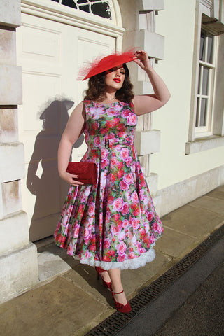 Elegant dark haired woman wearing a wide brimmed hat and a pink floral dress standing on a pretty English street in high heels clutching a red handbug.