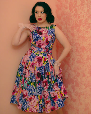 Dark haired woman with red lipstick standing against a wall wearing a bright floral print swing dress with a classic boatneck