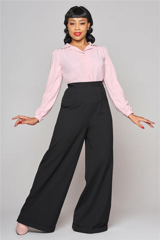 Tall slim woman with black curled hair standing with a happy smile wearing black high waisted wide leg trousers and a pink 40s style blouse.