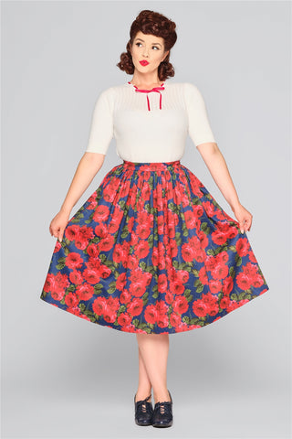 Elegant brunette woman holding out her skirt wearing a white knit top with a red ribbon detail neckline and a navy swing skirt with red roses print