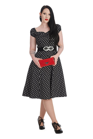 Dark haired rockabilly woman wearing a black and white polka dot dress and a statement belt with a red clutch and high heels