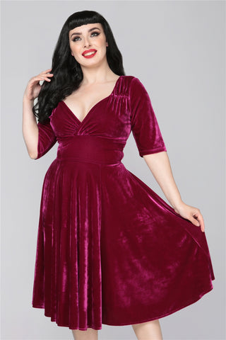 Woman with long black hair and red lipstick wearing a 50s style mid length deep burgundy dress with 3/4 length sleeves
