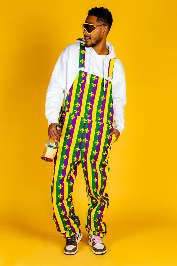 Man in Mardi Gras overalls holding a bottle, wearing sneakers and sunglasses, close-up of shoe and hand with bottle.