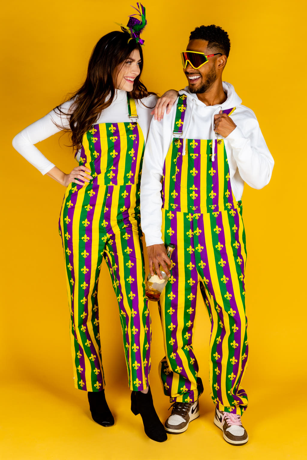 A man and woman in Mardi Gras overalls, ready for a day of celebration with beignets in hand.