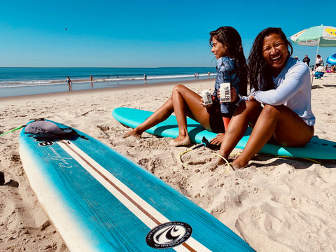 Two girls holding mini Boxed Water cartons on surfboard in sand