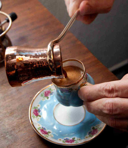 Unfiltered Turkish coffee being poured from a brass cezve, into a small ceramic mug. A saucer sits on a wooden table below.