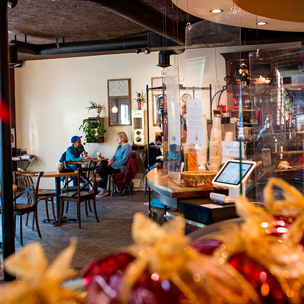 Two people sitting in a small cafe (the dining area of San Francisco Coffee). They each have a coffee and are having a discussion. In the foreground are images of wrapped loaves of bread for the holiday with bows and red decorative paper.