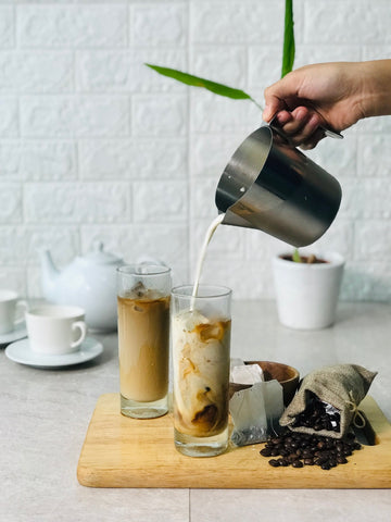 White background with a person pouring cream into a glass of coffee that sits on a cutting board