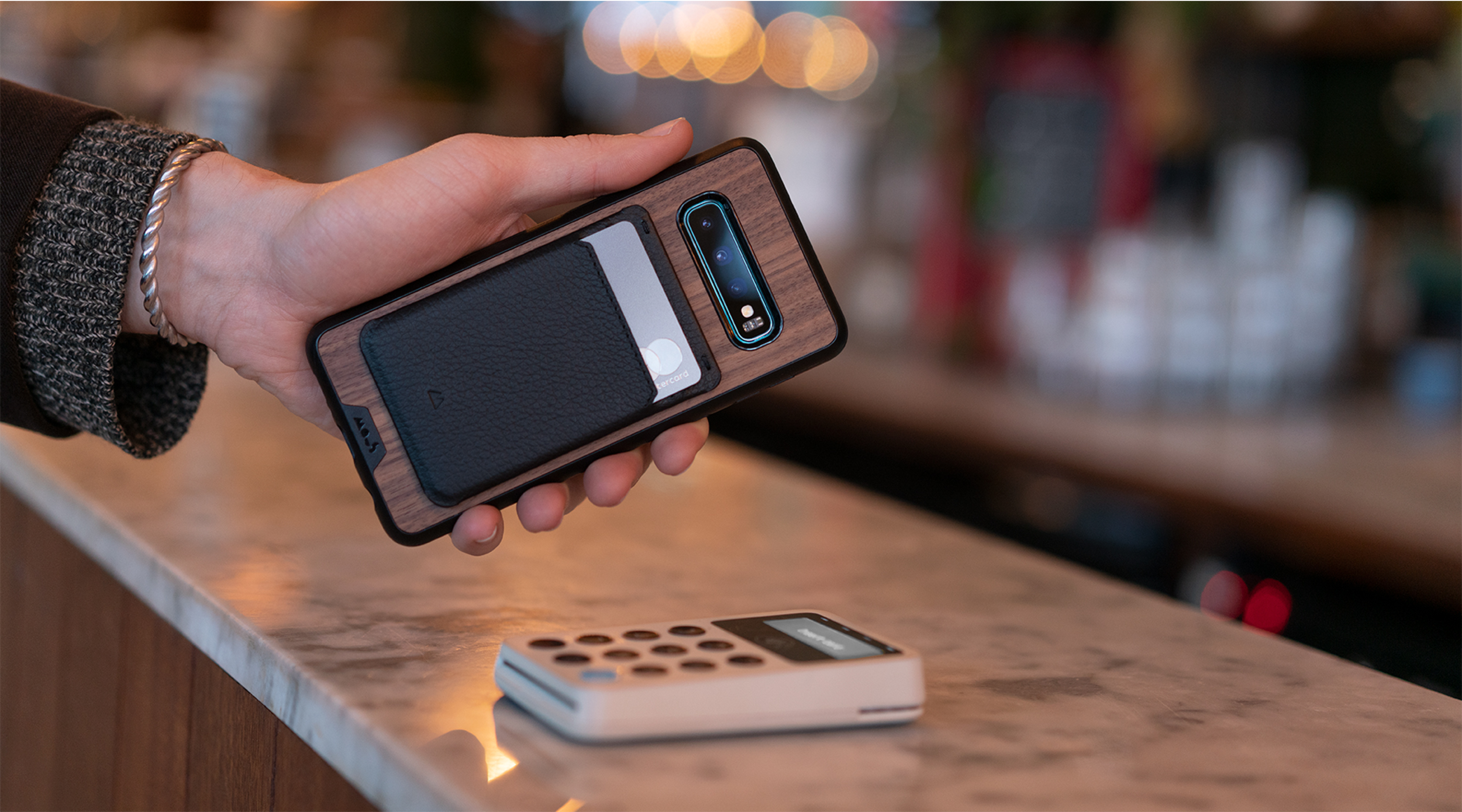 Person holding a walnut Samsung phone case with a phone card wallet above a card reader in a coffee shop