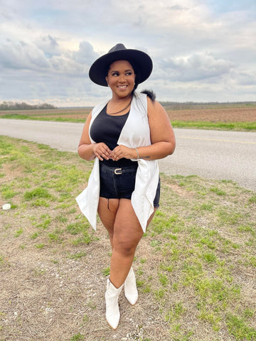Morgan B. Styles is standing in a dirt road wearing black gold chain fedora hat, black top, black ripped shorts, white vest, and white cowboy boots