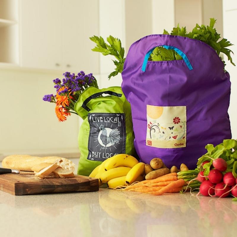 Onya Reusable Shopping bags holding items from the grocery store. They sit on a kitchen counter next to a bunch of bananas, potatoes, carrots, radishes and a partially sliced baguette on a cutting board. Available at Green Distributors.