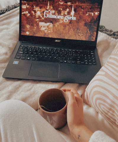 A girl holding a cup of tea while watching Gilmore Girls in ther cozy bed on a perfect fall day