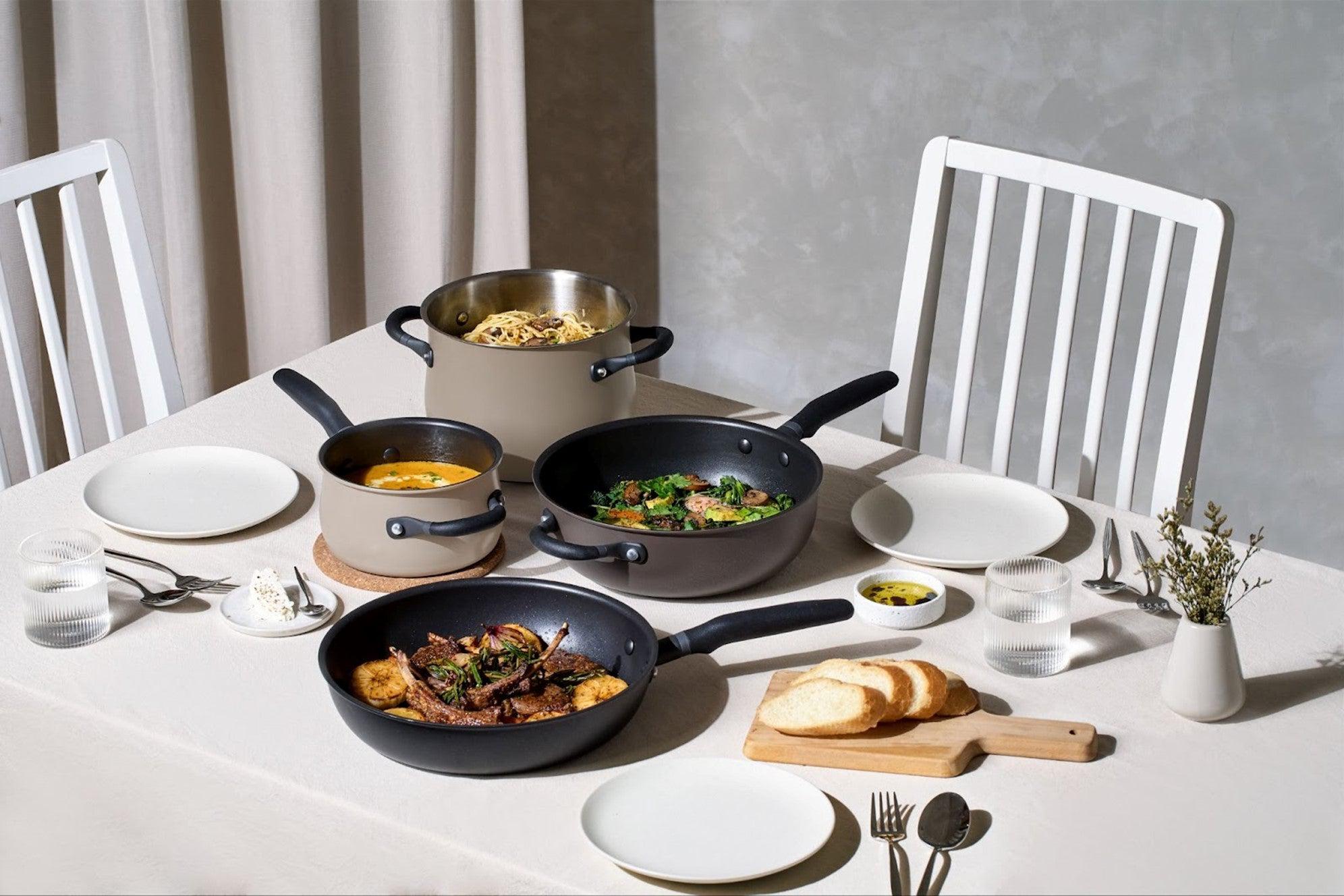The Meyer Cookware essential set in cinder and smoke colors, featuring a skillet pan, chef's pan, saucepan, and stockpot, beautifully presented with a delicious meal on a pristine white table setting